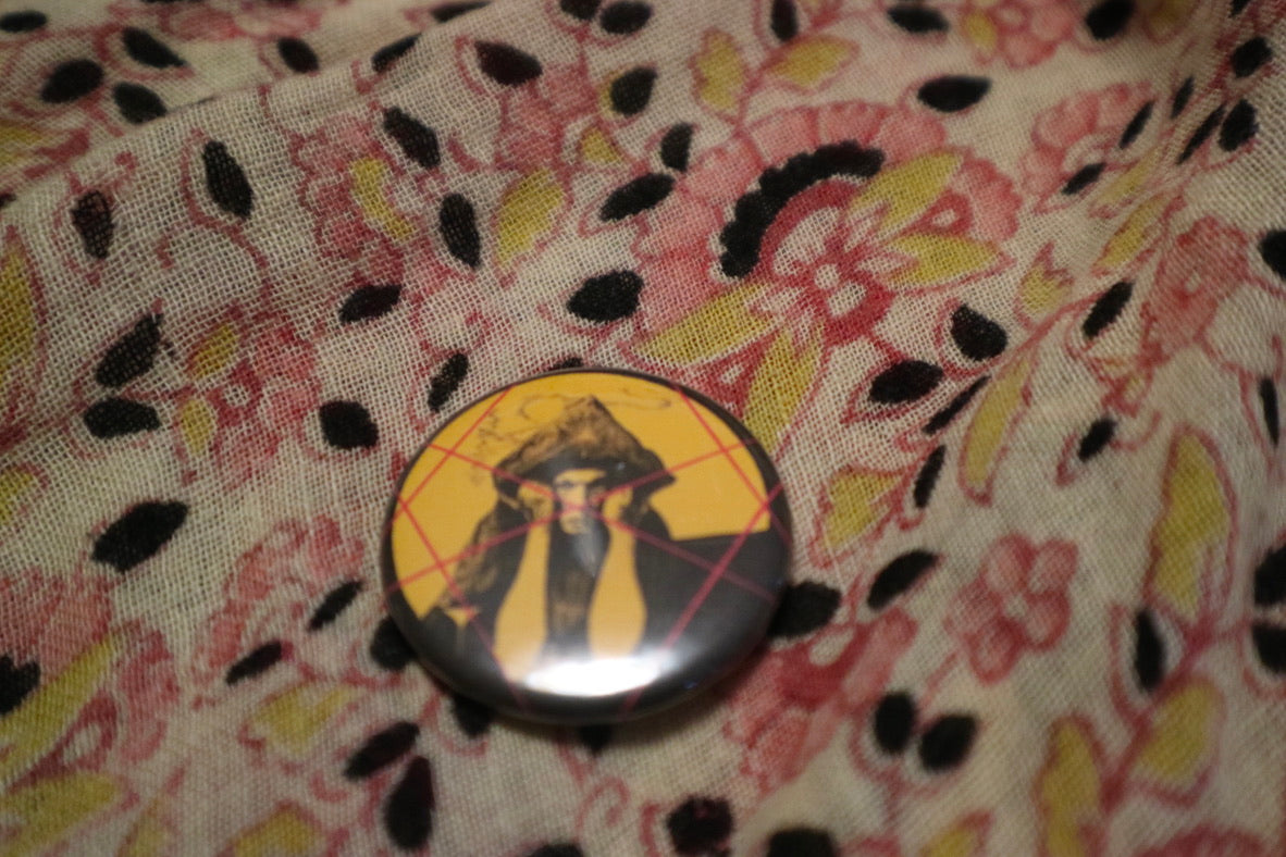 A yellow and red button badge of a portrait of Aleister Crowley on a paisley fabric